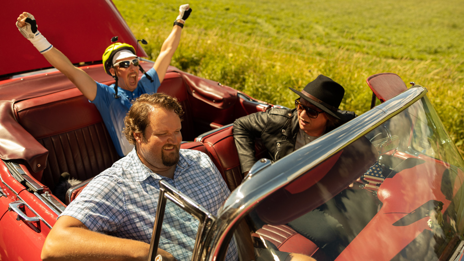 Kuvassa ovat punaisessa avoautossa etupenkillä ohjaajana Jaajo Linnoinmaa ja Sami Hedberg sekä takapenkillä Aku Hirviniemi kädet ojennettuina ylöspäin.  Kuva on otettu kesällä.