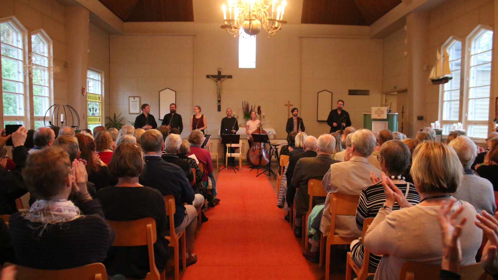 Kuvassa on konsertin päätös, jossa Klamilan kirkon etuosassa soittajat ottavat yleisön aplodeja vastaan.  Etualalla on yleisöä ja keskellä on käytävä.