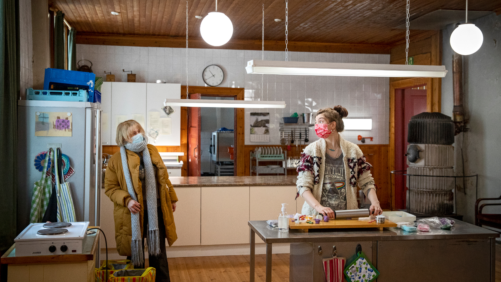 Kuvassa on vasemmalla päällysvaatteissa ja maskissa Leea Klemola ja oikealla leipomassa Emma Kilpimaa.  Kuva on keittiöstä.