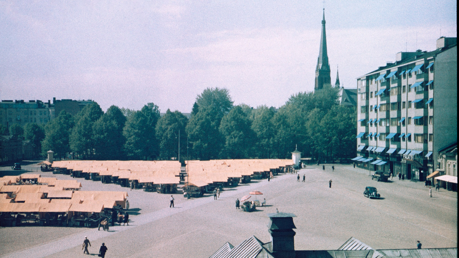 Kuvassa on Punaisenlähteen tori Viipurissa ja se on täynnä myyntikojujen telttakattoja ja taustalla näkyy kirkon torni.  Oikealla on kerrostalo.  Autoja näkyy vain kaksi.
