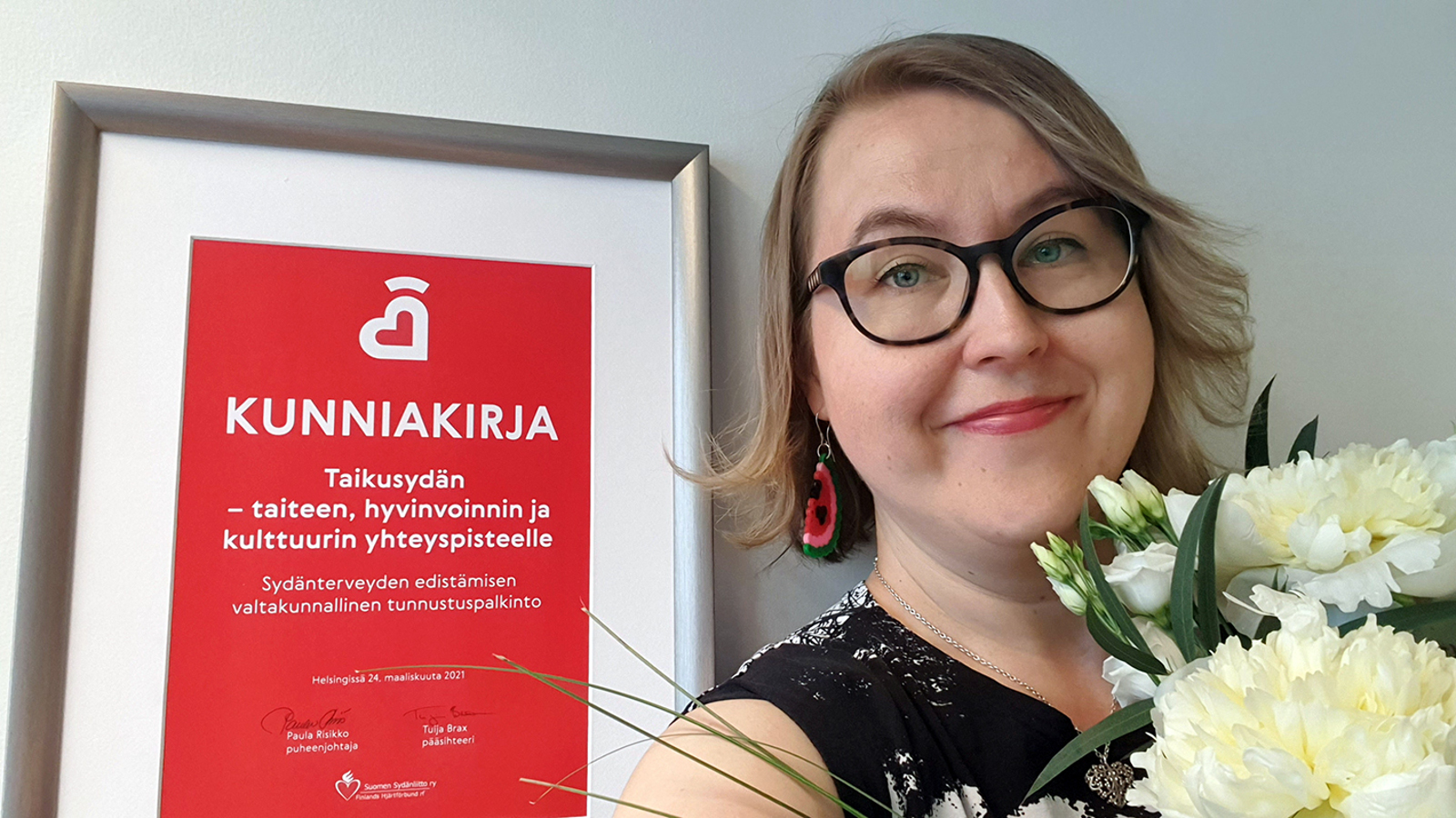 Kuvassa on vasemmalla Taikusydämen kunniakirja ja oikealla on Anna-Mari Rosenlöfin kasvokuva kukat kädessään.