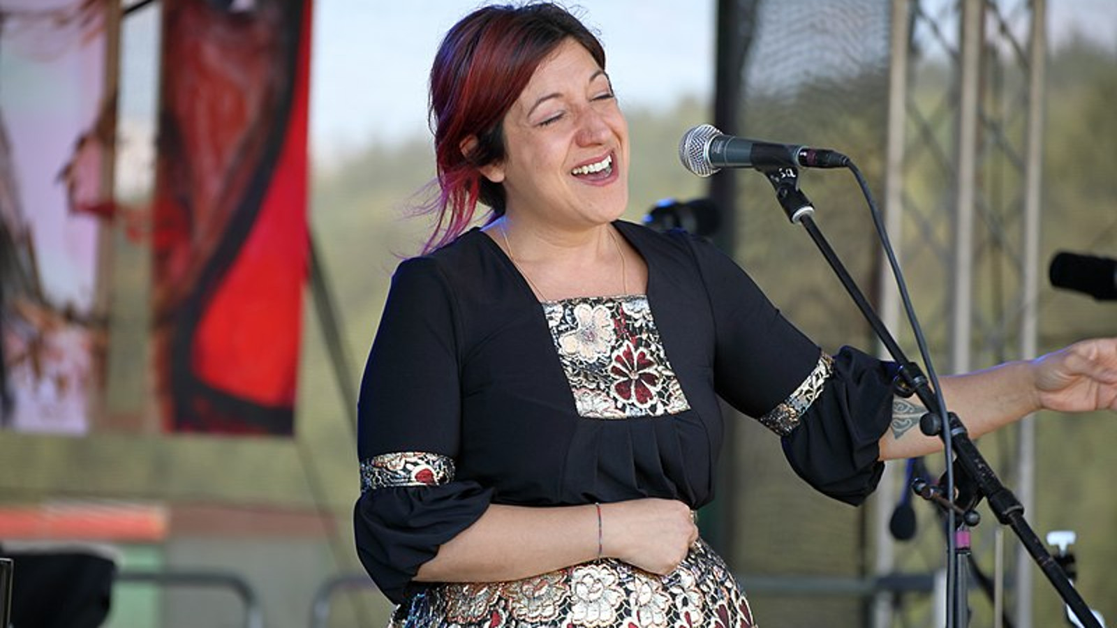 Kuvassa on Maria Mazzotta esiintymässä mikrofoni edessään.  Hänellä on kukallinen mekko päällään.