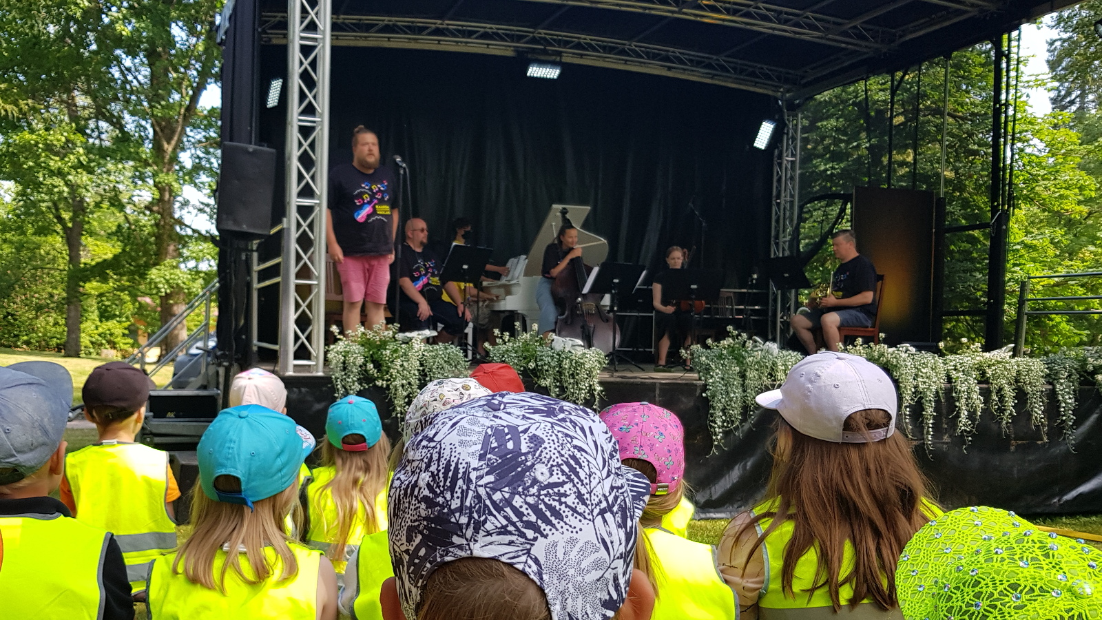 Kuvassa ovat etualalla huomioliiveissä ja lippiksissä takaapäin näkyvät päiväkotilapset ja taka-alalla korkeammalla näyttämö, jossa on soittajia sekä vasemmalla seisomassa laulaja Erik Rousi.