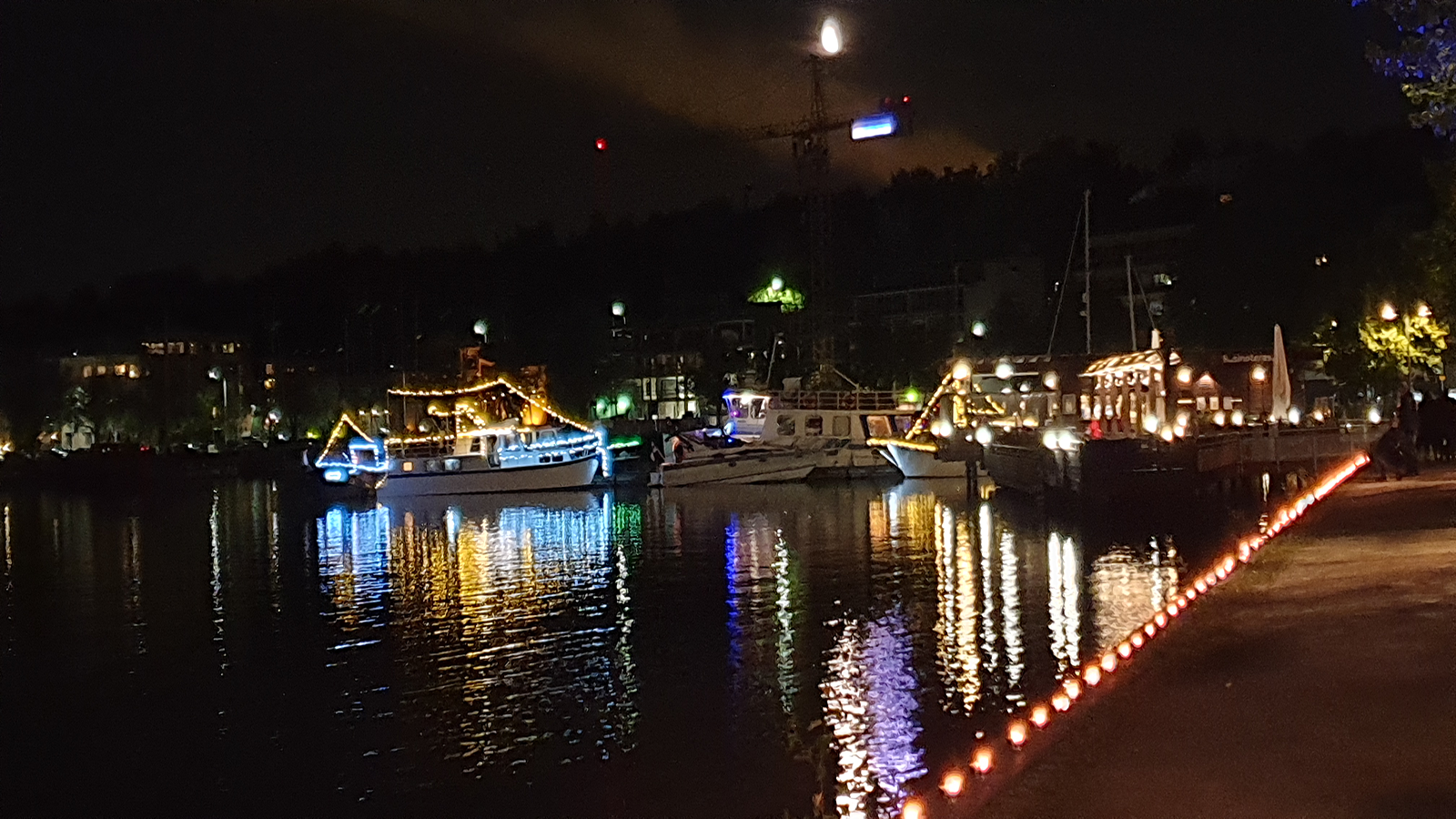 Kuvassa on satamassa oleva valaistuja veneitä ja rantaan penkalle sijoitettuja valoja.   
