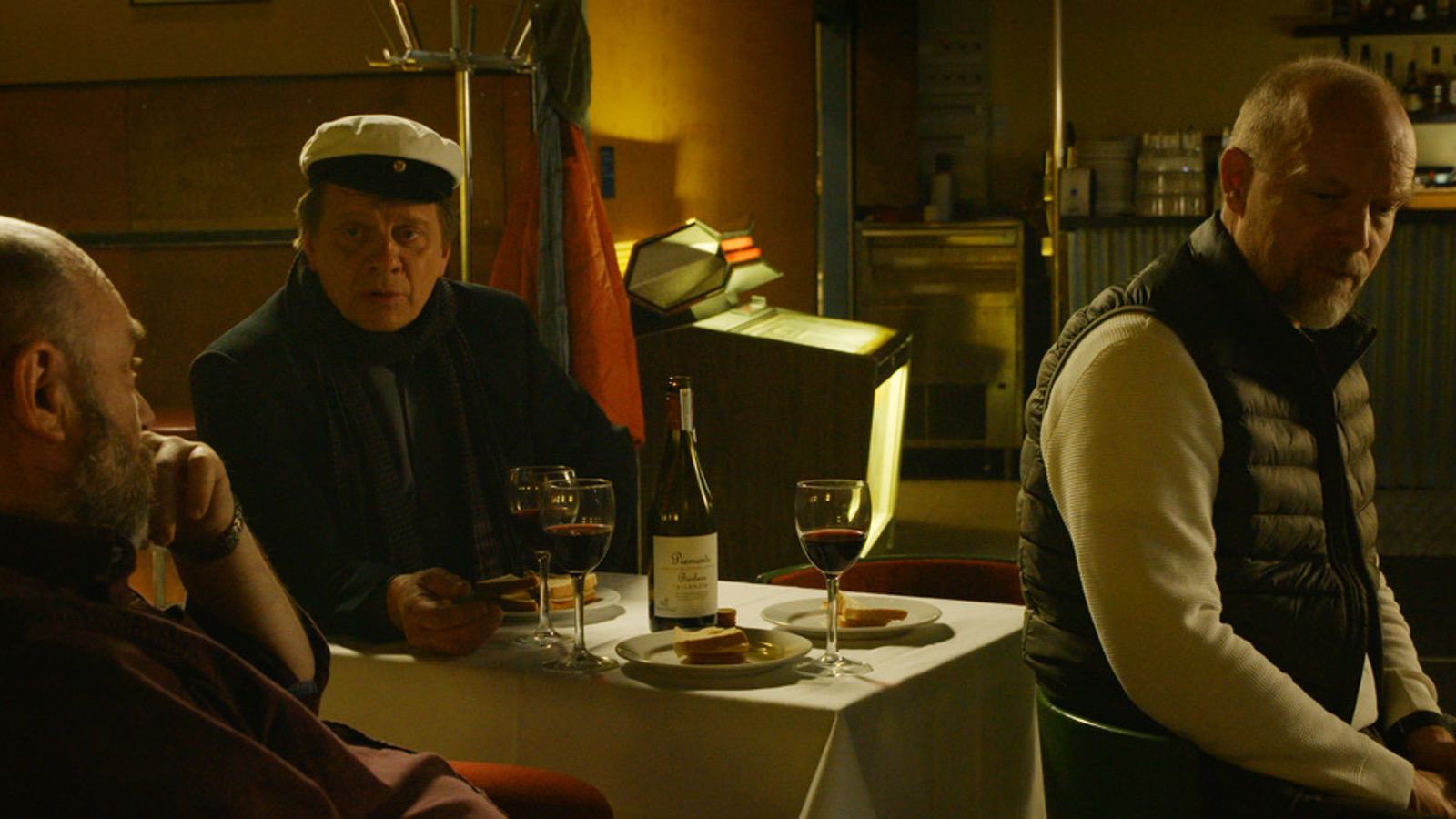 Kuvassa on hämärä baari ja sen pöydässä istuu kaksi ylioppilaslakissa olevaa miestä ja yksi oikealla oleva vinosti tuolillaan.