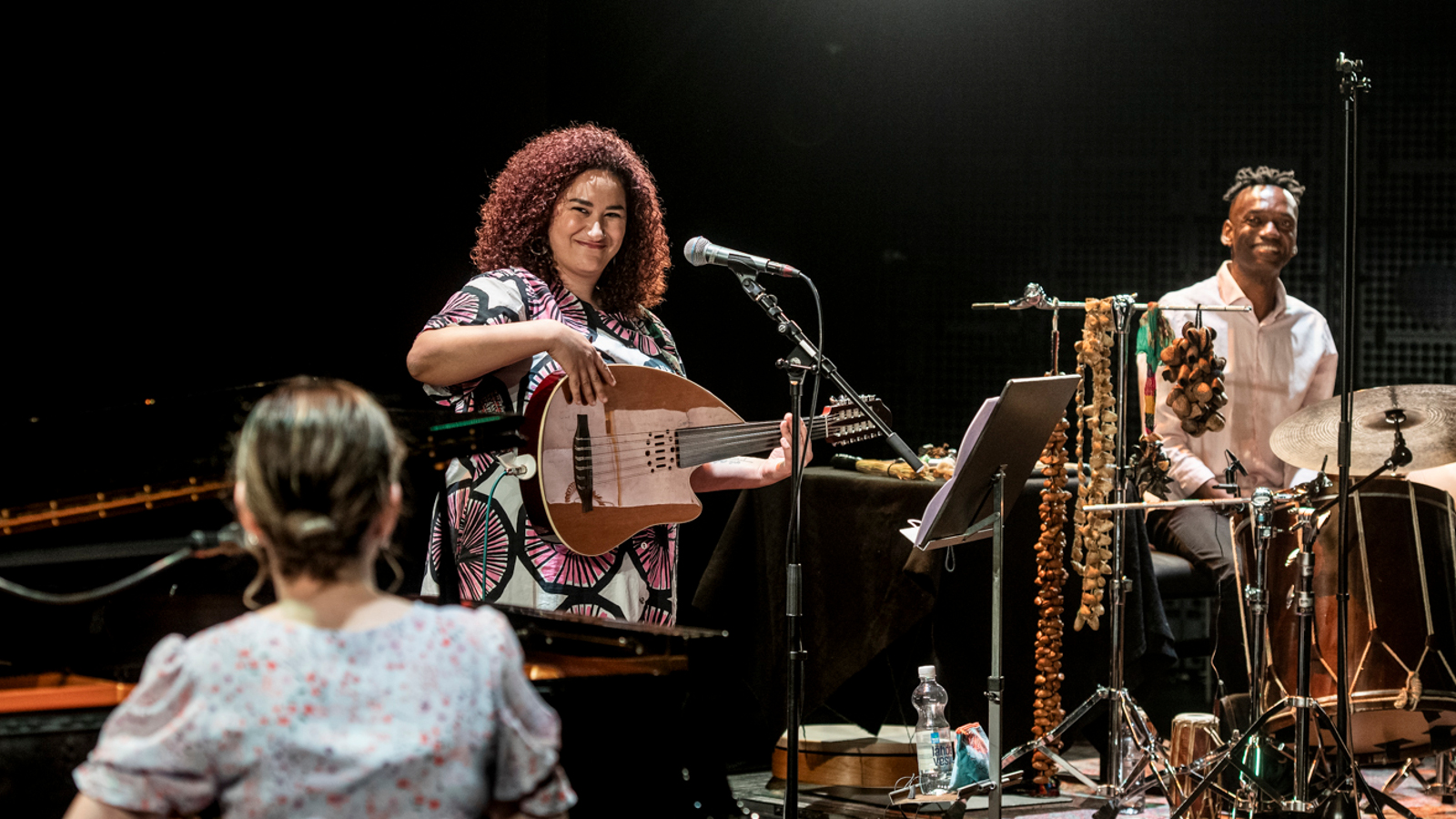Kuvassa on esiintymislavalla soittamassa tummaa taustaa vasten kitaraan kiharatukkainen nainen ja hänen vieressään mies haitari jaloissaan.  Vasemmalla etualalla selin nainen soittamassa pianoa.