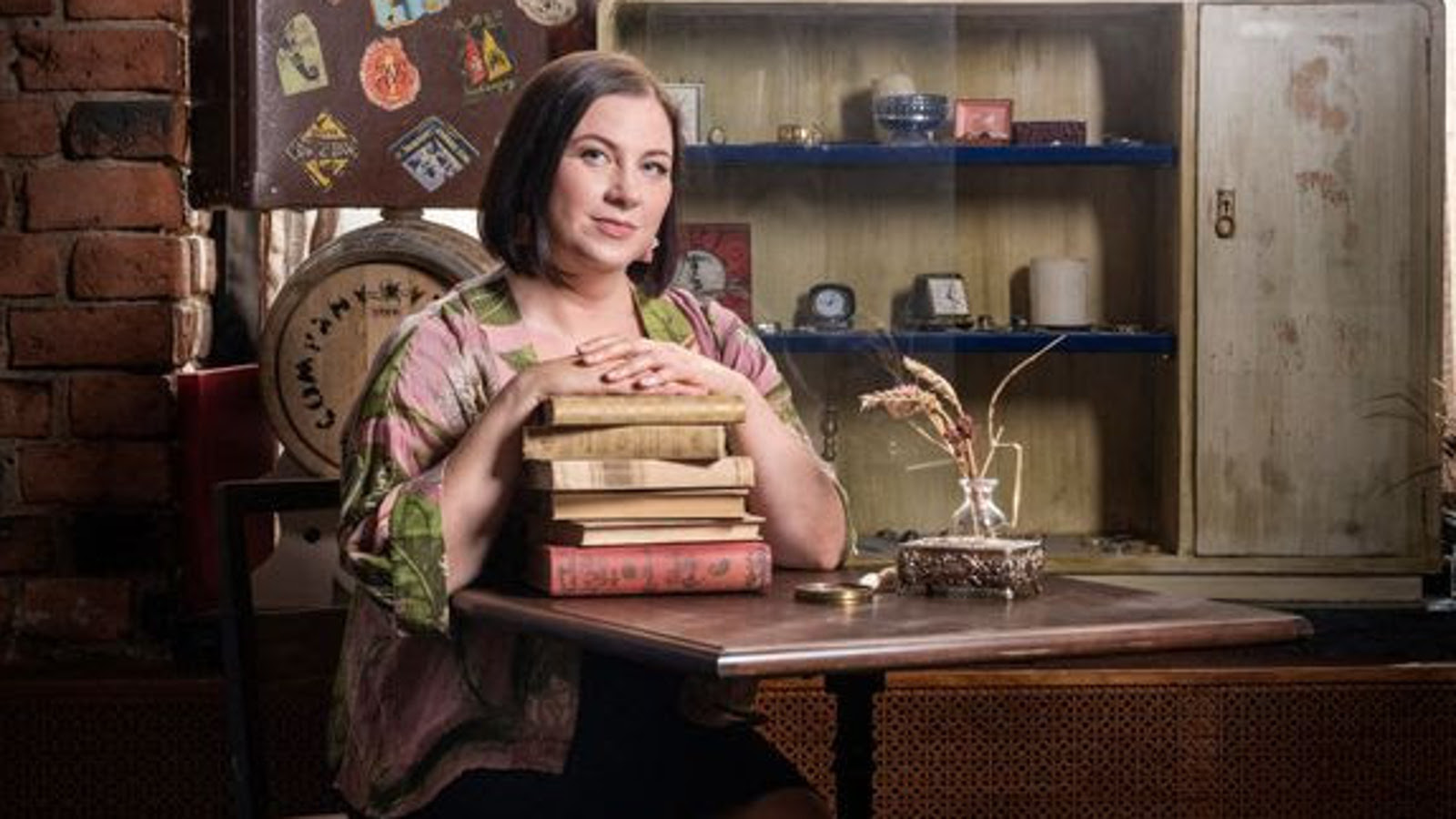 Kuvassa on Janica Brander istumassa pöydän ääressä kirjapino käsiensä alla. Taustalla näkyy kirjahylly ja kuvan sävy on ruskea.