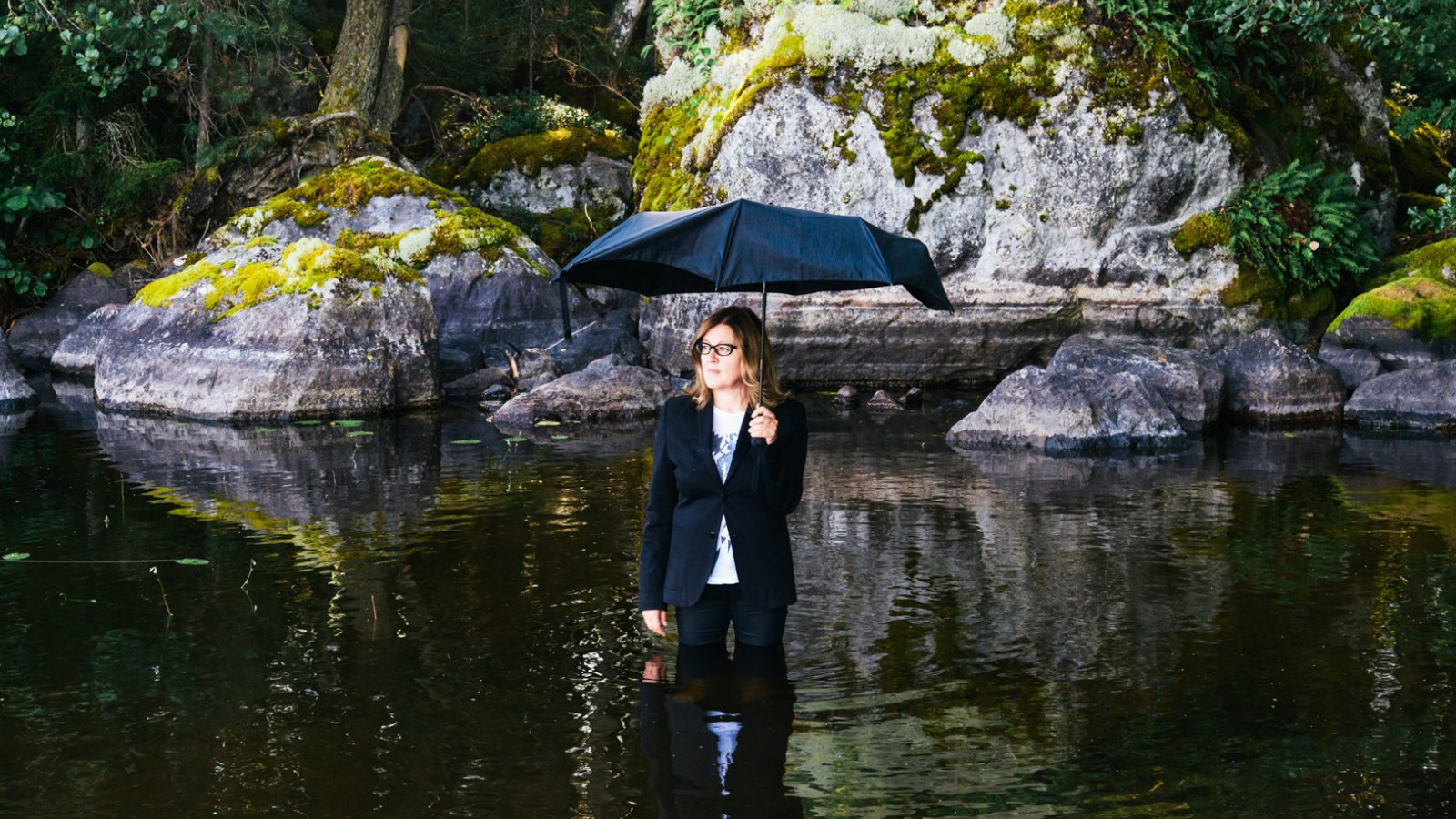 Kuvassa on Eija-Liisa Ahtila, joka seisoo tummassa vedessä polvia myöten tummassa pleiserissä ja pitkissä housuissa.  Hän pitää sateenvarjoa kädessään. Takana on kalliota.