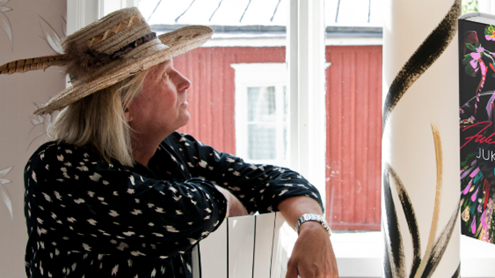 Kuvassa on Jukka Rintala istumassa sivuttain ja katsomassa ikkunasta ulos ja sieltä näkyy puna-valkoinen rakennus.  Rintalalla on lierihattu päässään ja hatussa on sulka.