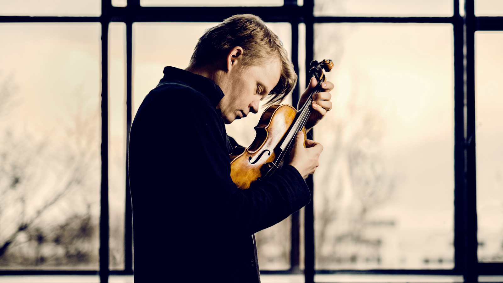Kuvassa on Pekka Kuusisto sivukuvassa viulu kädessään ja takana on isoja lasiristikoita.