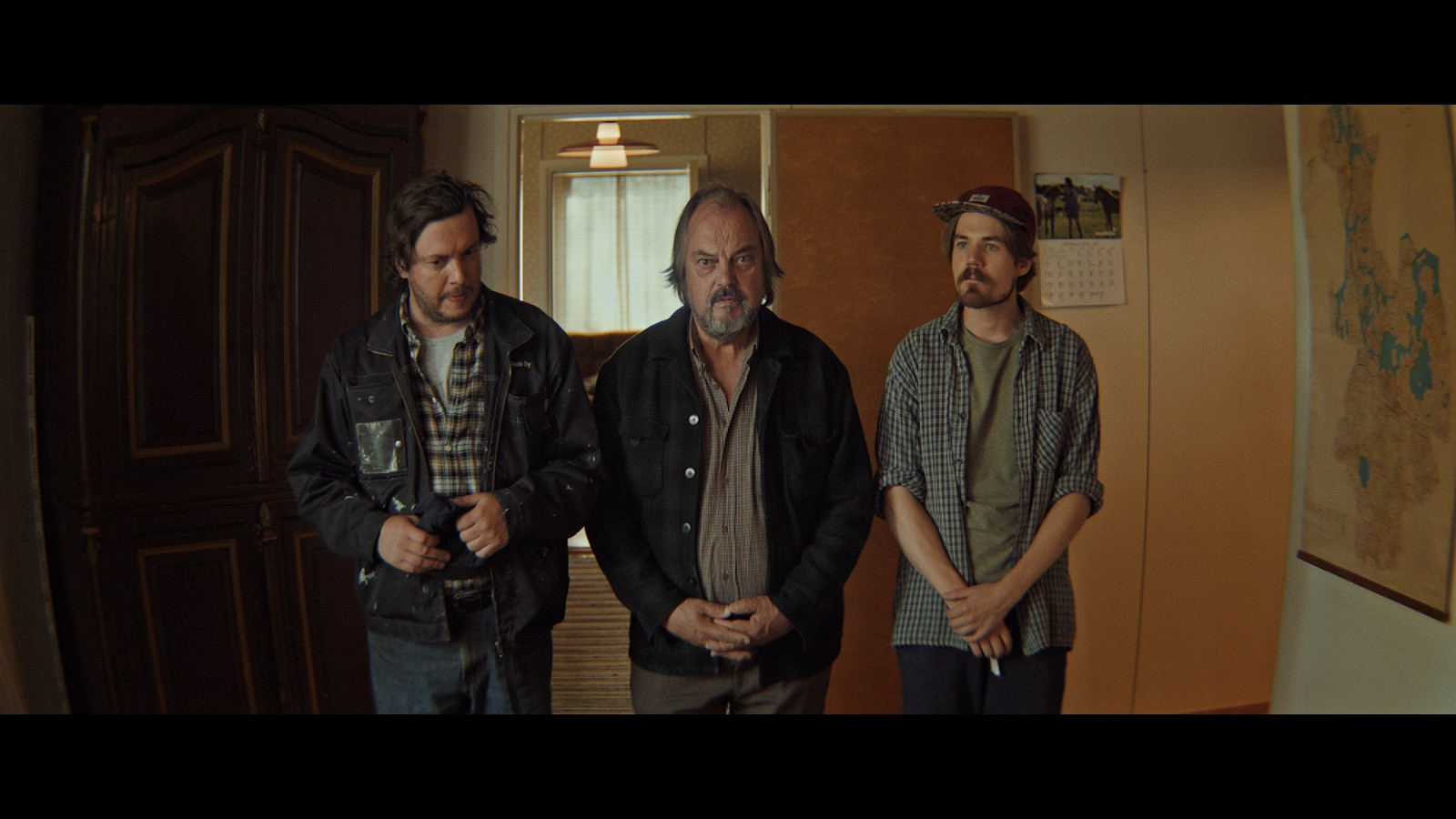 Kuvassa on kolme miesnäyttelijää seisomassa keskellä huonetta.  Kuvan sävy on tumma.
