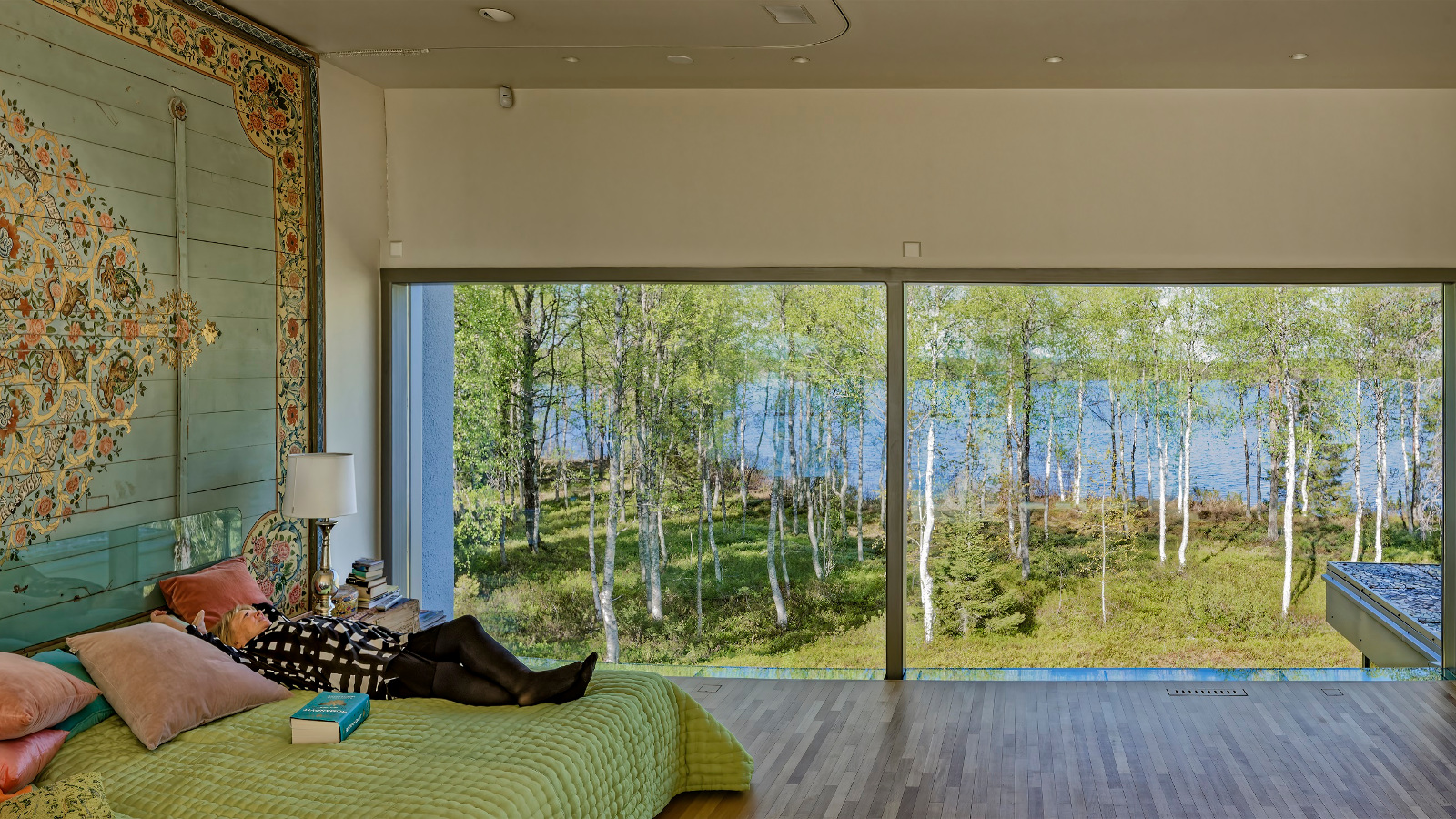 Kuvassa on Anu Pentikin koti, jossa on vihertävät sävyt. Anu Pentik makaa sängyllä ja katsoo ulo isoista ikkunoista.  Ikkunasta näkyy koivumetsää. 