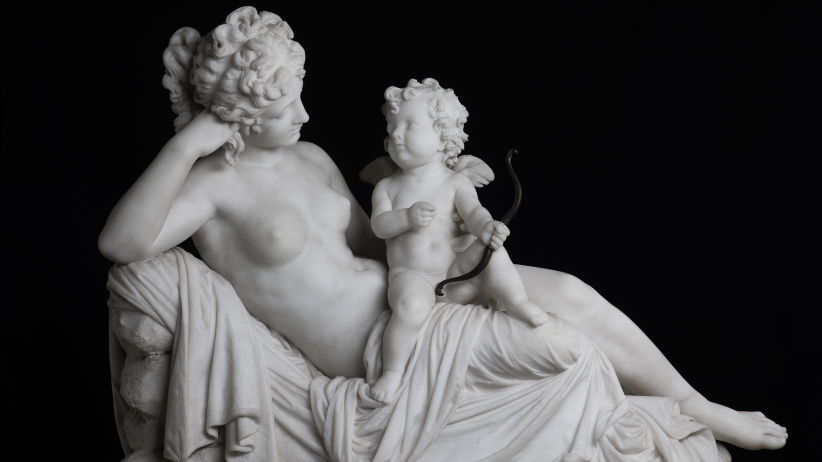 Kuvassa on Johannes Takasen vaalea veistos, jossa nainen istuu Amor nuolineen hänen sylissään