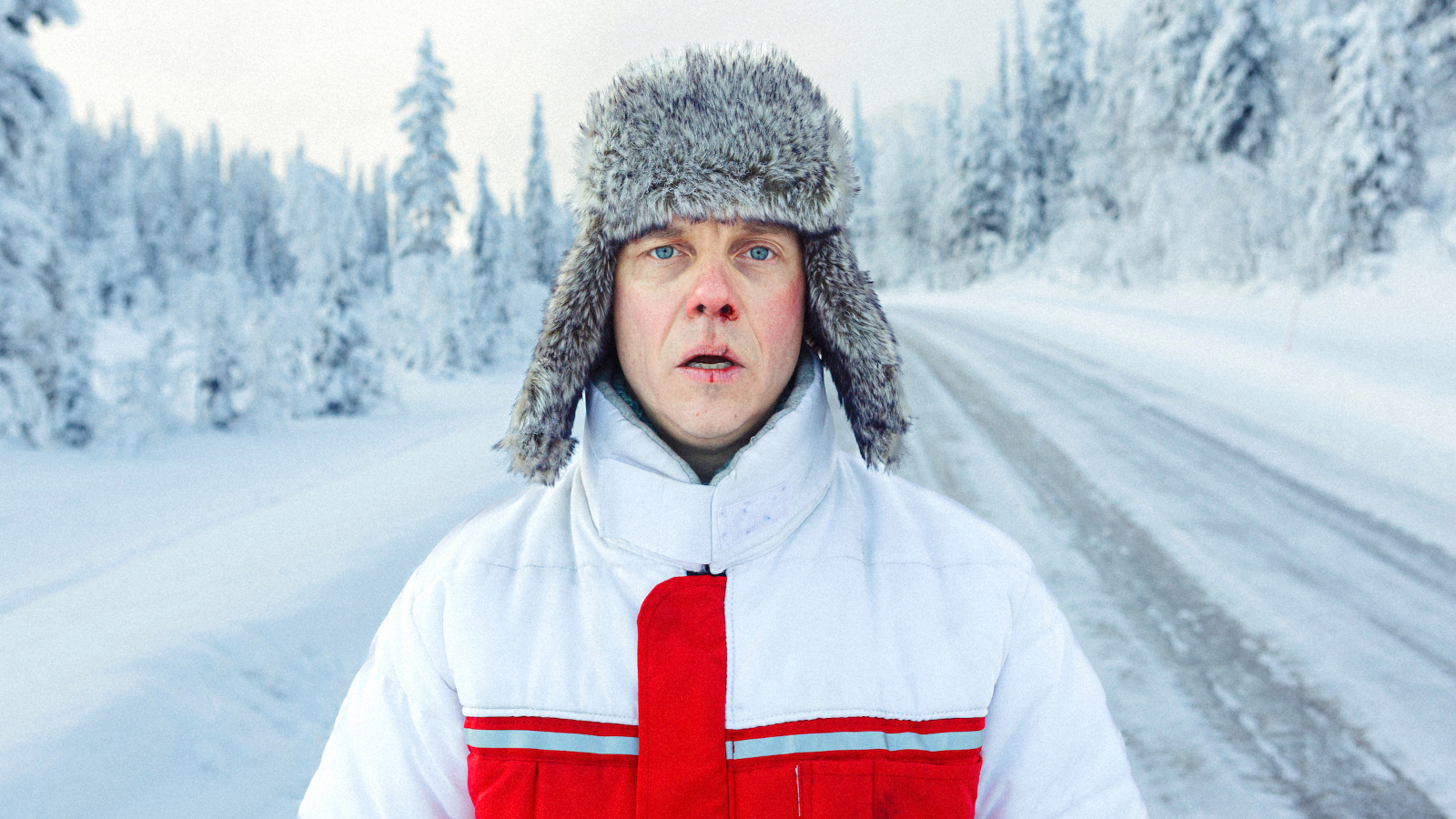 Kuvassa on Jarkko Lahti lumisessa maisemassa talvisella maantiellä.  Hänellä on valkoinen toppatakki, jossa on punainen ristikuvio. 
