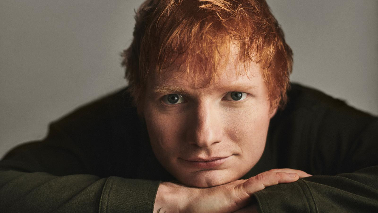 Kuvassa on Ed Sheeran, siten että hänellä on kädet pöydällä ja kasvot käsien päällä.