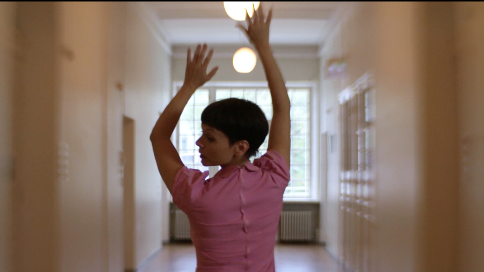 Kuvassa on Elli Isokoski tanssimassa käytävässä kädet ylhäällä ja hän on selin kasvot sivuttain vasemmalle. Kuva on puolivartalokuva.