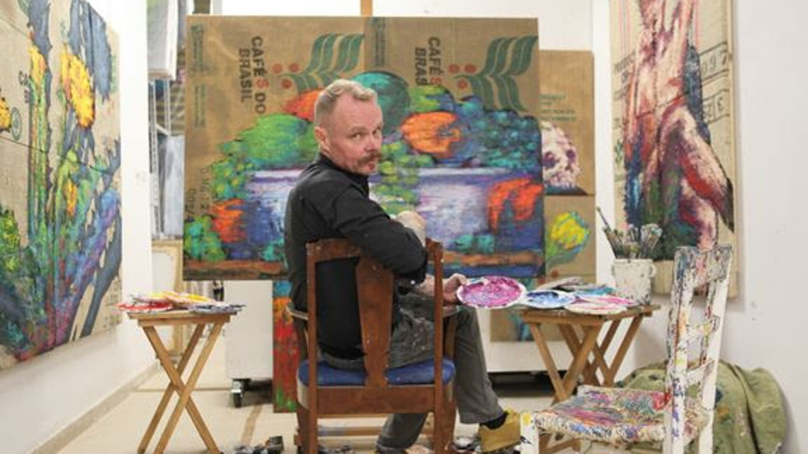 Kuvassa on Hannu Palosuo on istumassa ja tekee taideteosta.  Hän on kääntyneenä katsojaan päin.  Seinillä hänen ympärillään on taideteoksia.