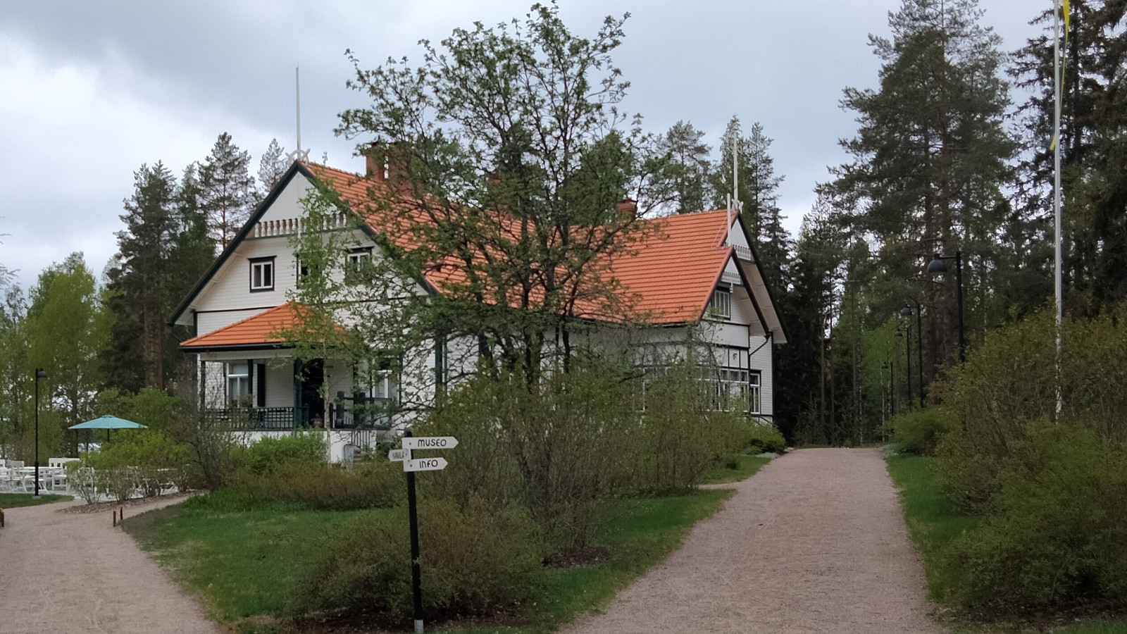 Kuvassa on Kotkaniemen päärakennus, joka näkyy vinosti ja etualalla on pihatiet muovaten nurmikon v-mallisesti taloa kohti.  Talo on puiden katveessa ja on väriltään vaalea vihrein reunalaudoituksin.