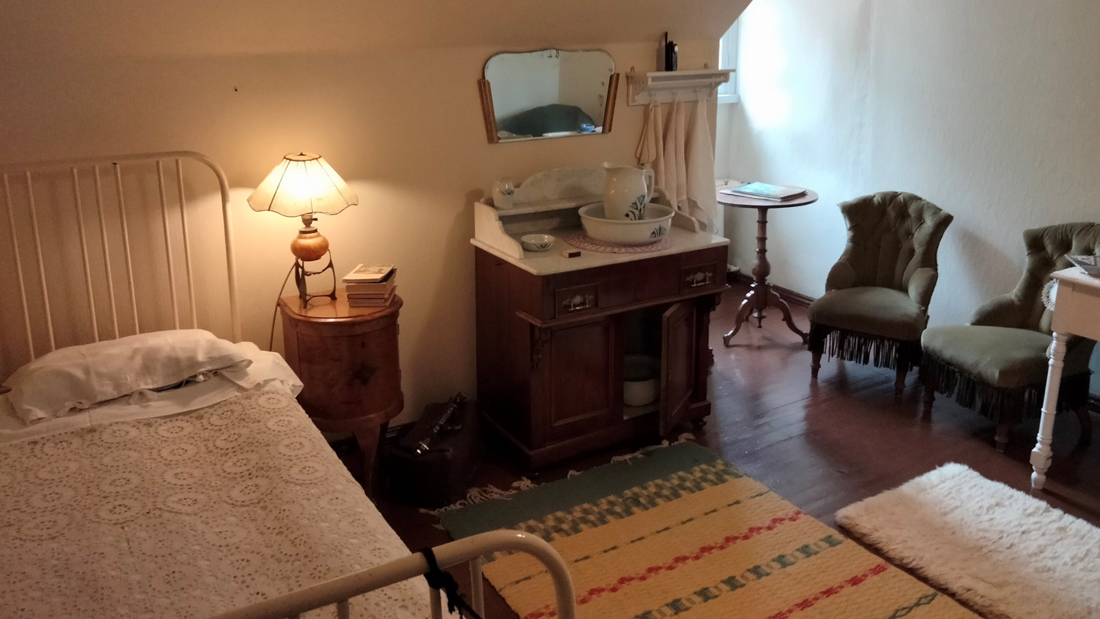 Kuvassa on vinokattoinen ullakkohuone, jossa on vasemmalla metallipäätyinen sänky, jossa on vaalea peite.  Vieressä on puinen yöpöytä, jonka päällä on lamppu ja vieressä pieni piironki.