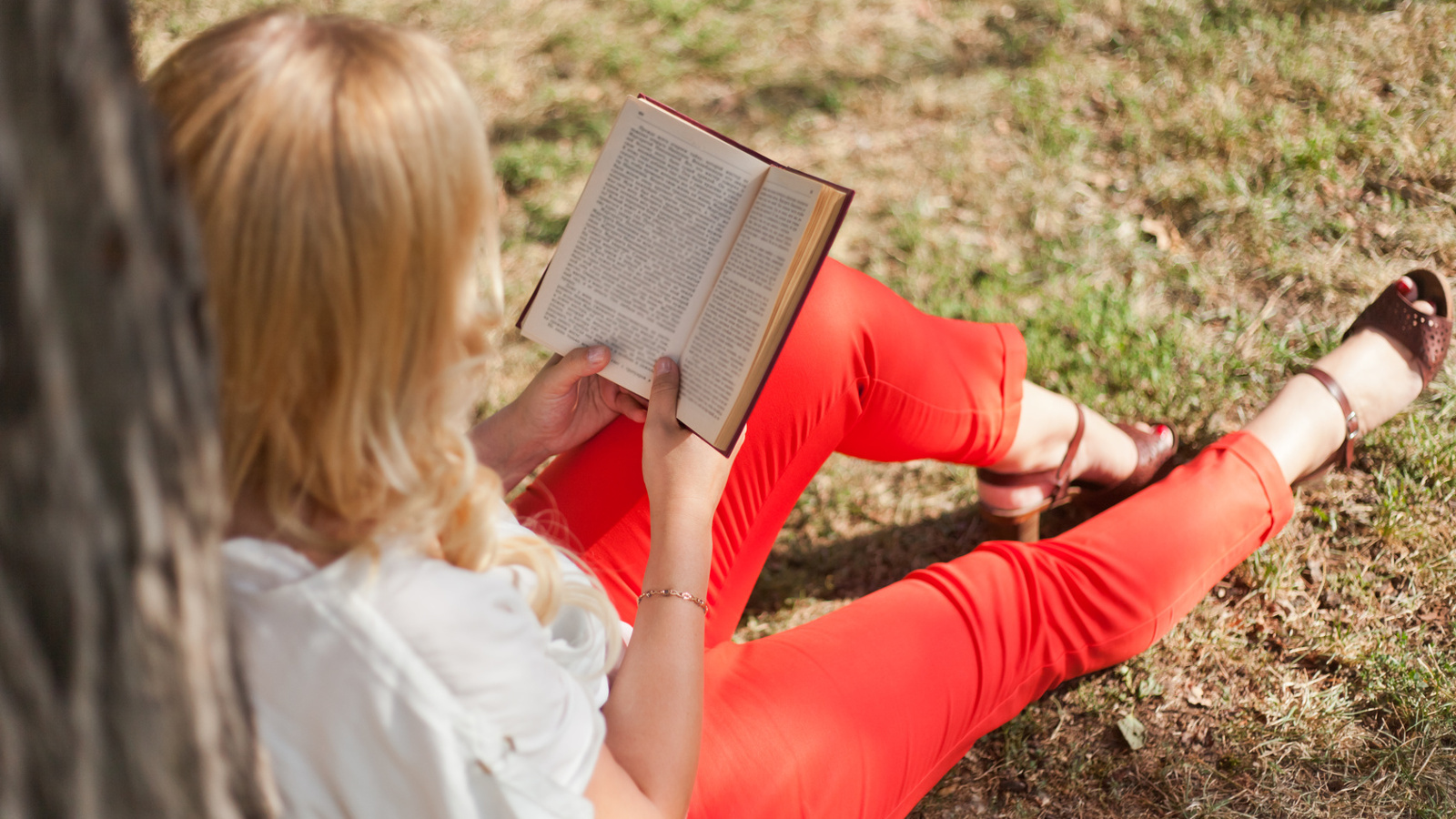 Kuvassa on nainen kuvattuna yläviistosta ja hän lukee istuvassa asennossa nurmikolla kirjaa.  Hänellä on jalassaan punaiset housut.