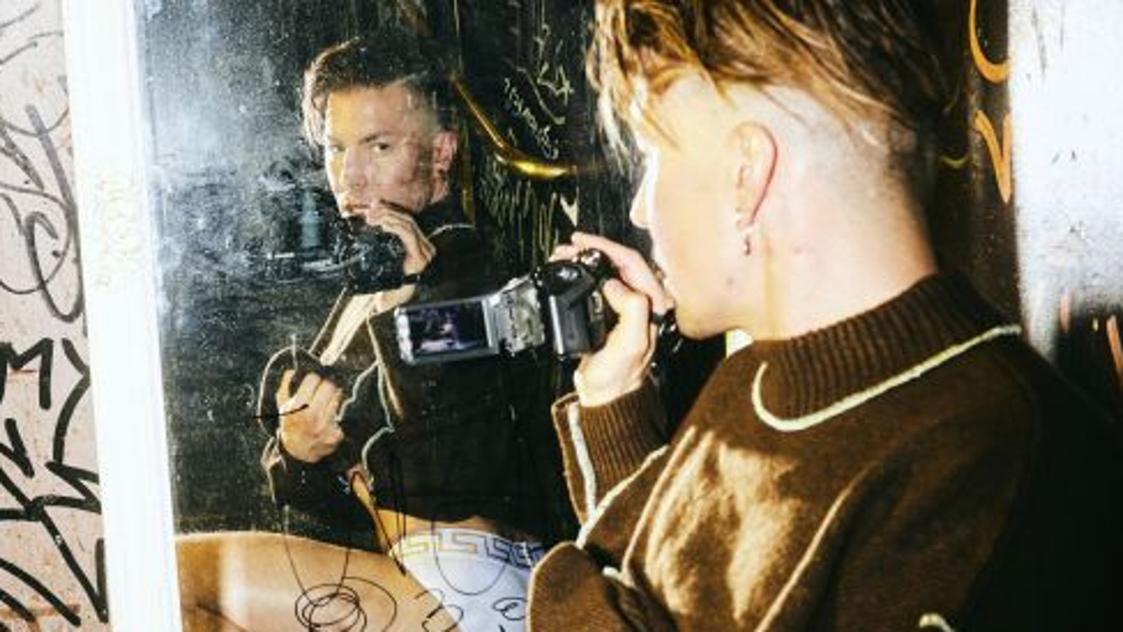 Kuvassa on Antti Tuisku, joka katsoo peilistä itseään ja hänellä on kamera kädessään. Kuvassa on ruskeita graafisia elementtejä.