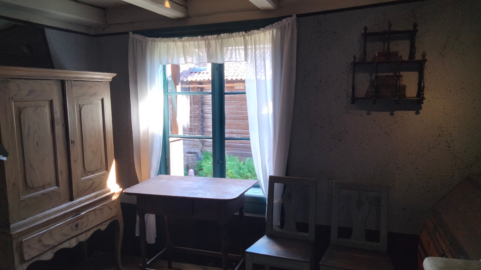 Kuvassa on huone, josta näkyy kadulle ja vasemmalla seinällä on puinen kaappi.  Ikkunan edessä on pöytä.