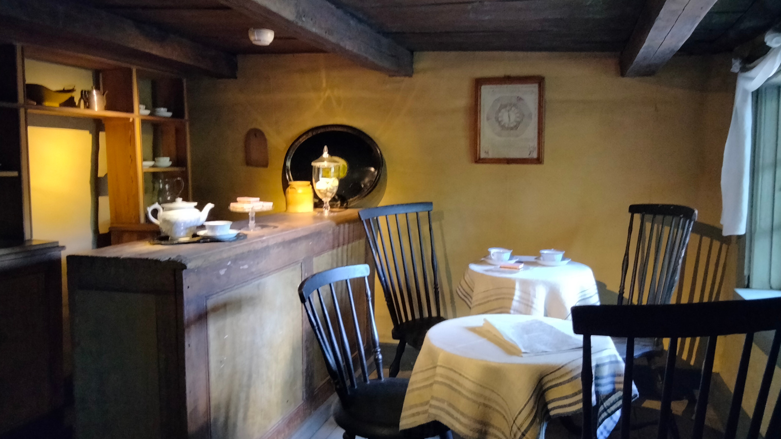 Kuvassa on kahviohuone, jossa on vasemmalla on palvelutiski, jossa on vanhanaikaisia astioita. Oikealla valkoisten pöytäliinojen pöytiä kaksi.