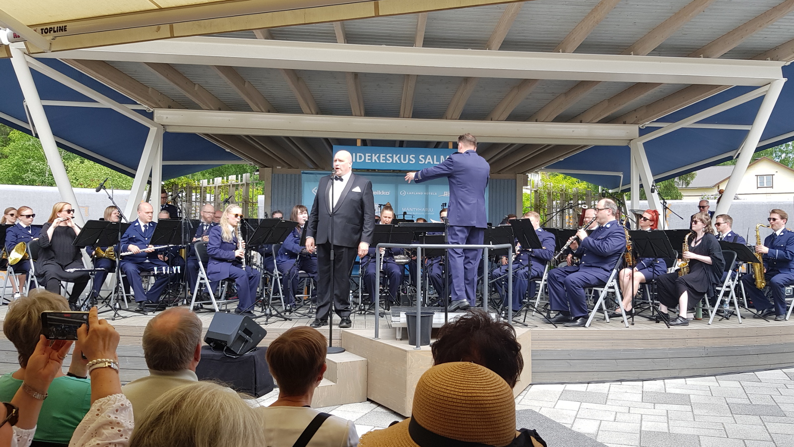 Kuvassa on Mika Pohjonen laulamassa Poliisisoittokunnan edessä. Taustalla poliisisoittokunta soittamassa sinisissä poliisipuvuissa.