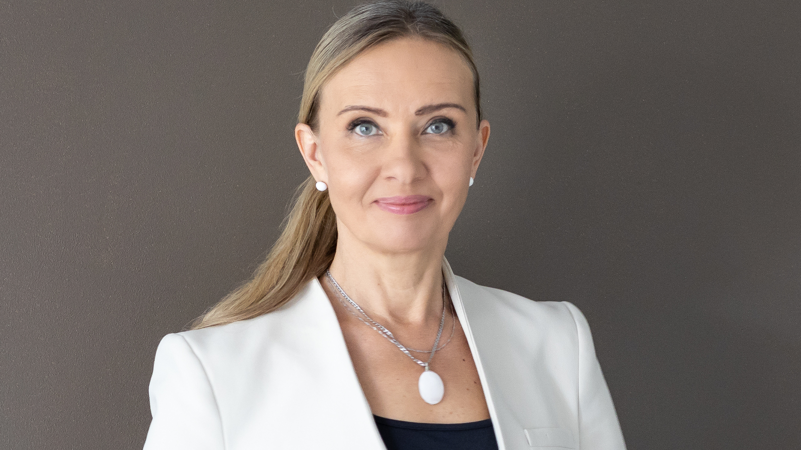 Kuvassa on Ilmi Villacís valkoisessa jakussa, mustassa  topissa ja kaulassaan hopeinen soikea kaulakoru. Hänellä on pitkät hiukset kammattuna taakse poninhännälle. Kuvan tausta on ruskea.