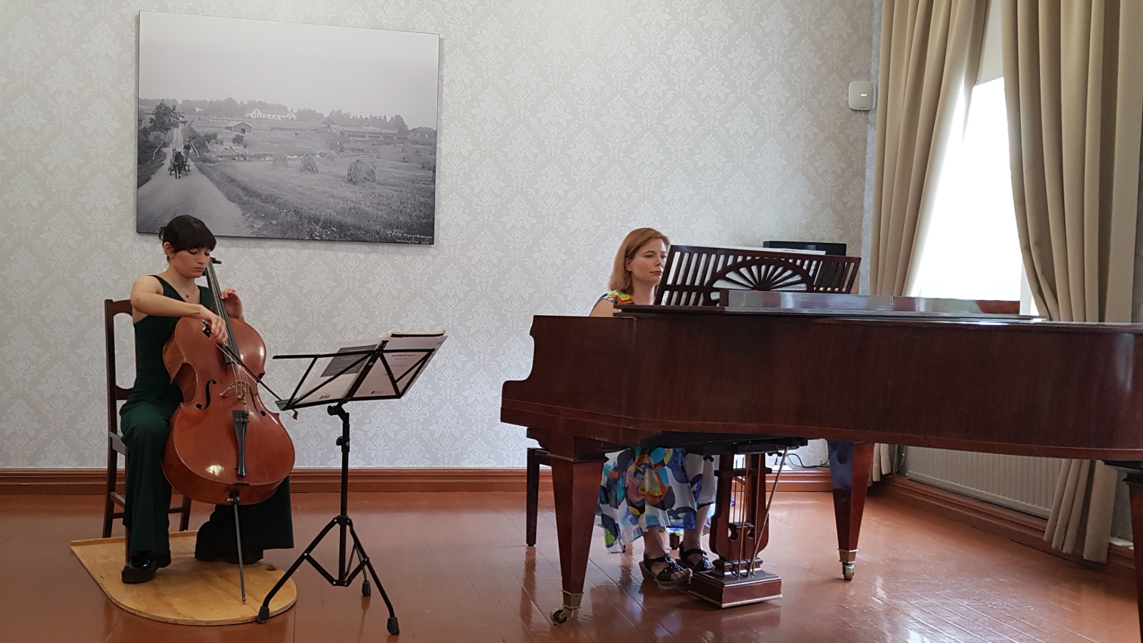 Kuvassa on Maria Morfin Venäläinen soittamassa selloa tumman vihreässä housuasussa ja oikealla Liisa Malmivaara säestämässä flyygelin ääressä. Taustalla on musta-valkoinen taulu vanhasta valokuvasta.