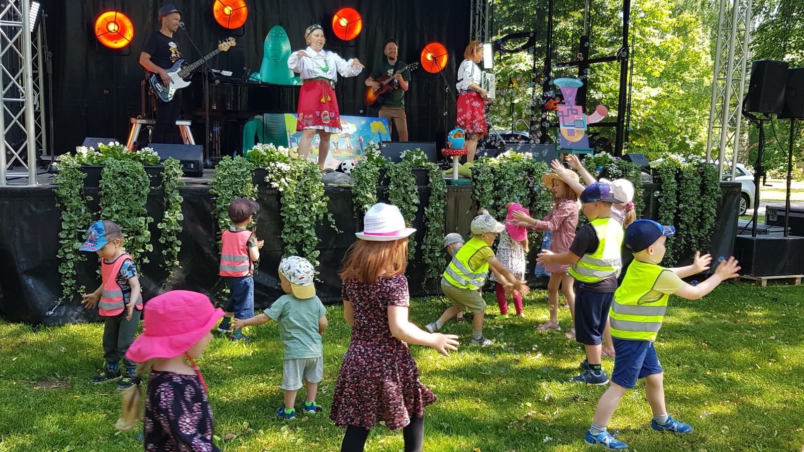 Kuvassa ovat Mimmien esiintyjät Pauliina Lerche ja Hannamari Vallila punaisissa puolihameissa ja taustalla bändin soittaja flyygelissä, basisti ja kitaristi. Puistonäyttämön edessä alhaalla nurmikolla on joukko lapsia leikkimässä.