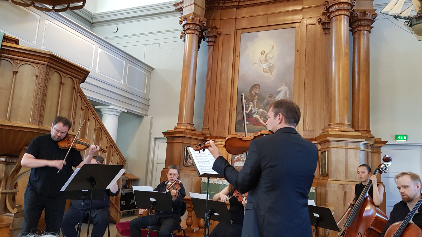Kuvassa ovat Petteri Iivonen ja Jukka Merjanen selin soittamassa viulua ja taustalla viulistit ja basson sekä sellon soittajat.  Kaikilla on mustat esiintymisasut.