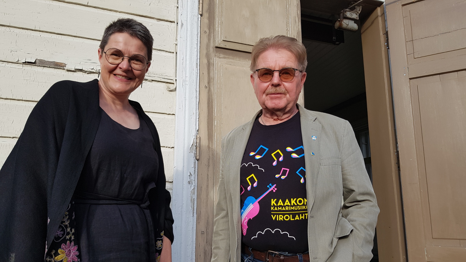 Kuvassa ovat Siru Ahopelto mustissa vaatteissa ja Risto Kouki kamarimusiikin t-paidassa. Taustalla on Virolahden kirkon vaaleata seinää ja ruskeaa kirkon ovea.