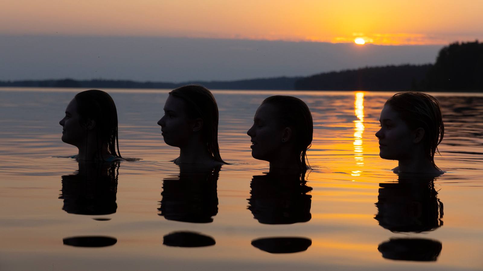 Kuvassa on auringonlaskun vesimaisemassa neljän pään silhuetit veden päällä.