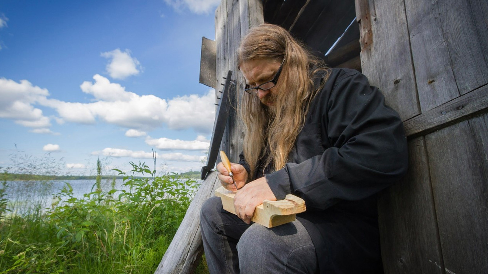 Kuvassa on Rauno Nieminen soittamassa pientä kannelta järven rannalla harmaan venevajan seinän vieressä. 