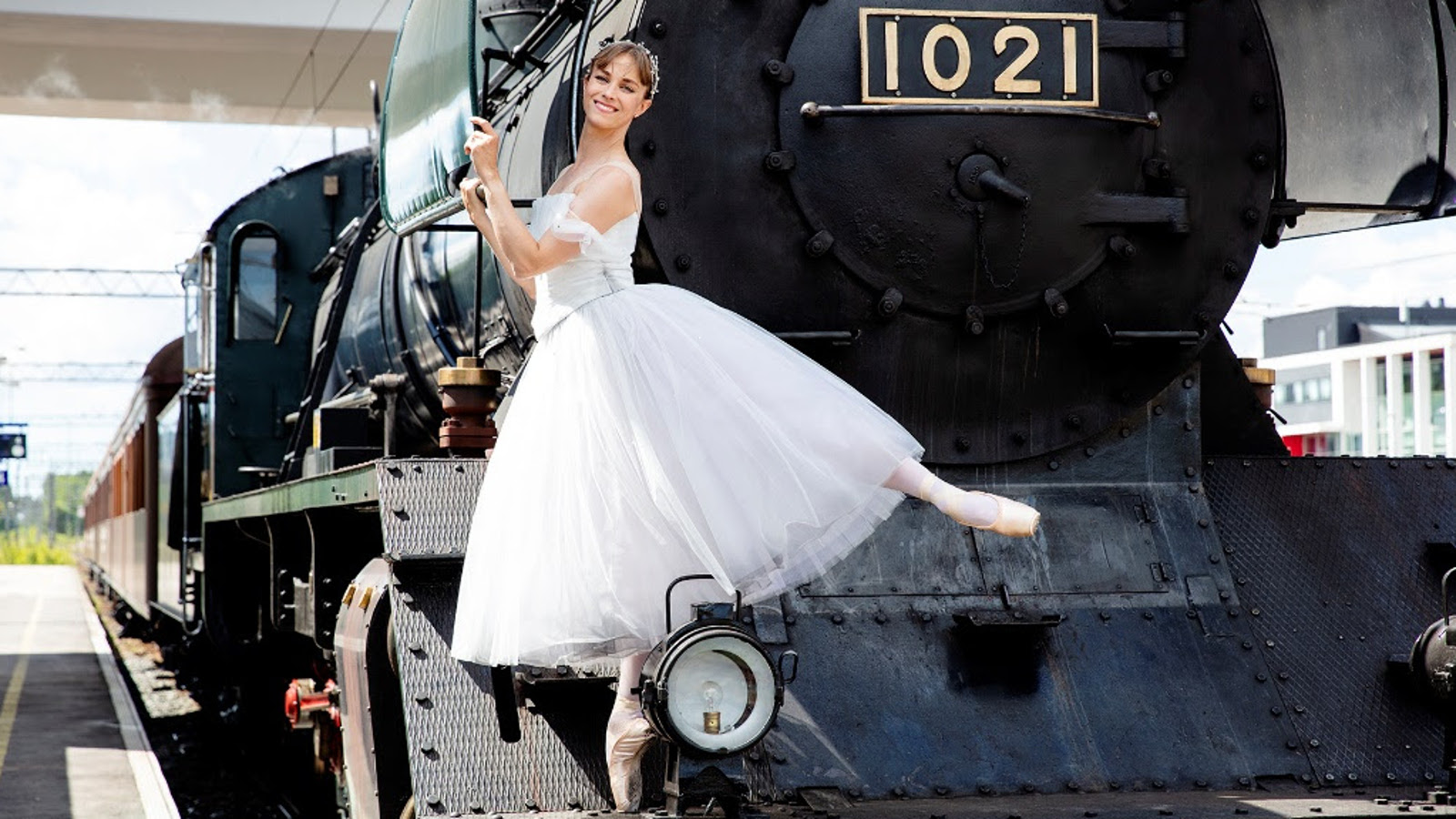 Kuvassa on ballerina valkoisessa tyllipuvussa toinen jalka ojennettuna taakse vanhan höyryveturin no 1021 nokalla.