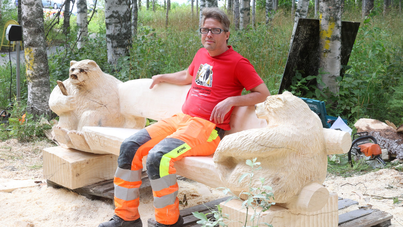 Kuvassa puupenkki, jossa on molemmissa päissä veistetyt karhun kuvat.  Penkillä istuu veistäjä, jolla punainen t-paita ja keltaiset pitkät housut.