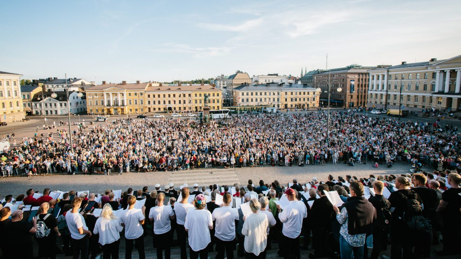 Kuvassa on edessä suurkuoro esiintymässä senaatintorilla yleisölle, joka seisoo torilla.