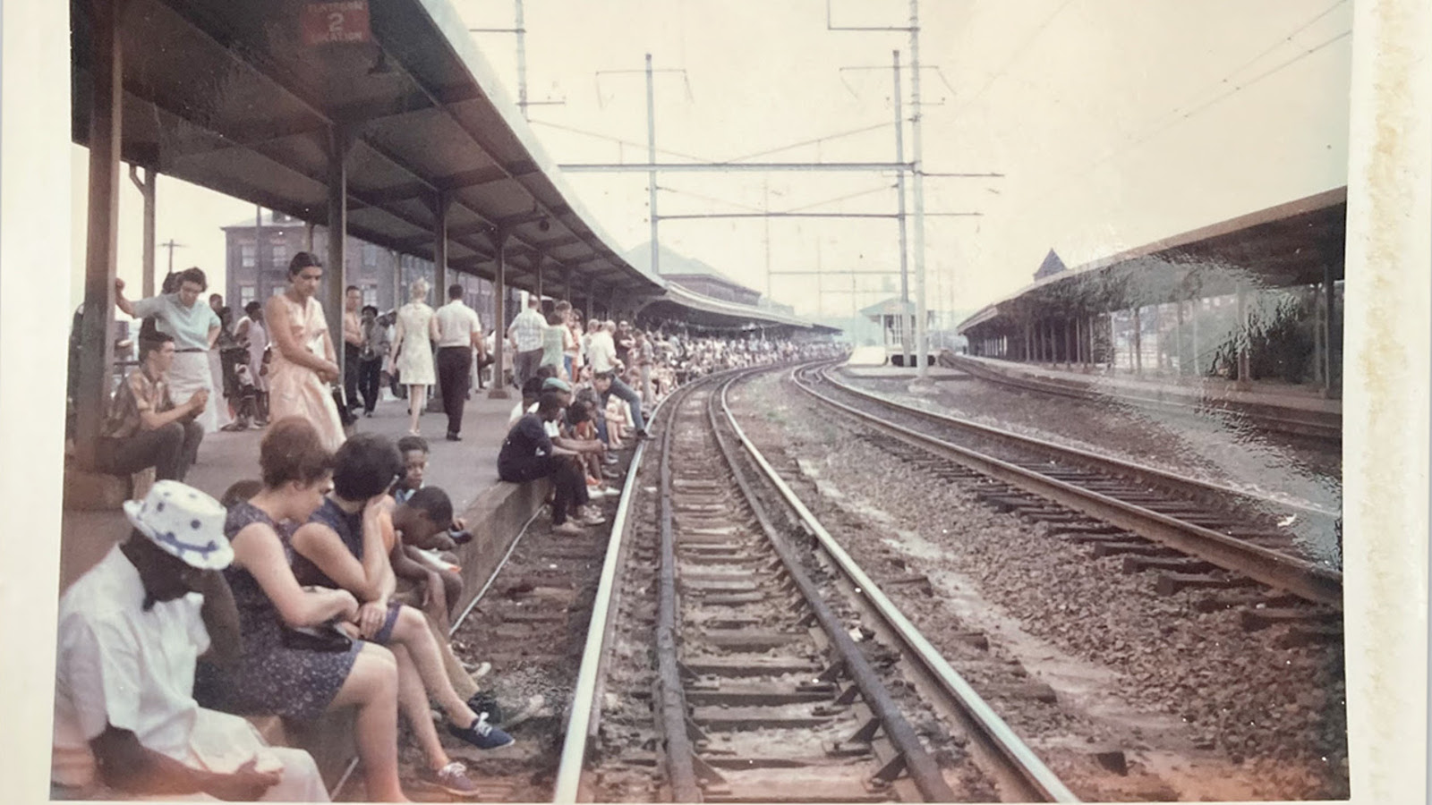 Kuvassa on ihmisiä istumassa ja seisomassa katoksen alla ja odottamassa junaa. Raiteet kaartuvat oikealle.