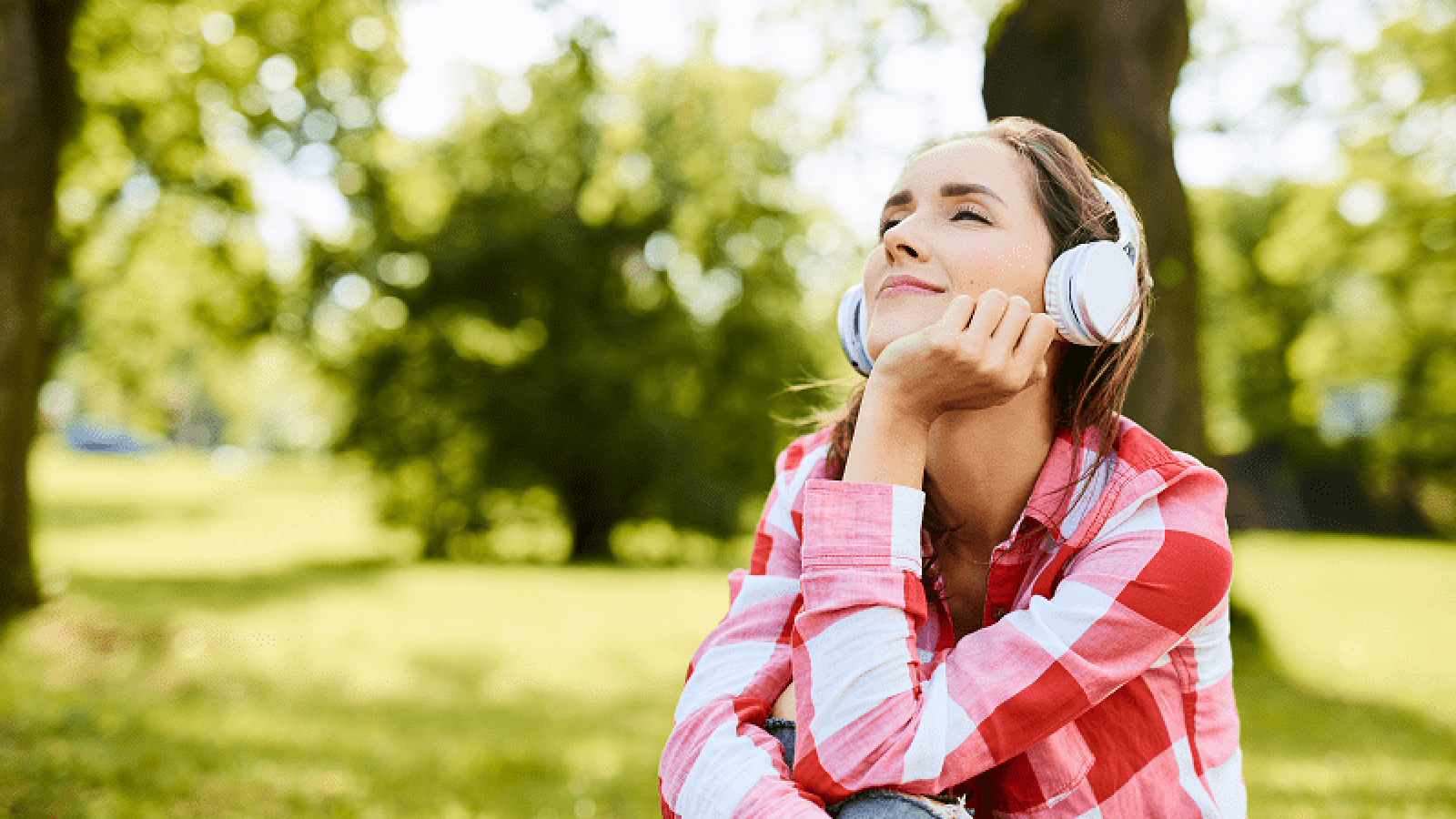 Kuvassa on musiikkia kuunteleva nuori nainen kuulokkeet päässään. Taustalla on vihreää puistoa. 