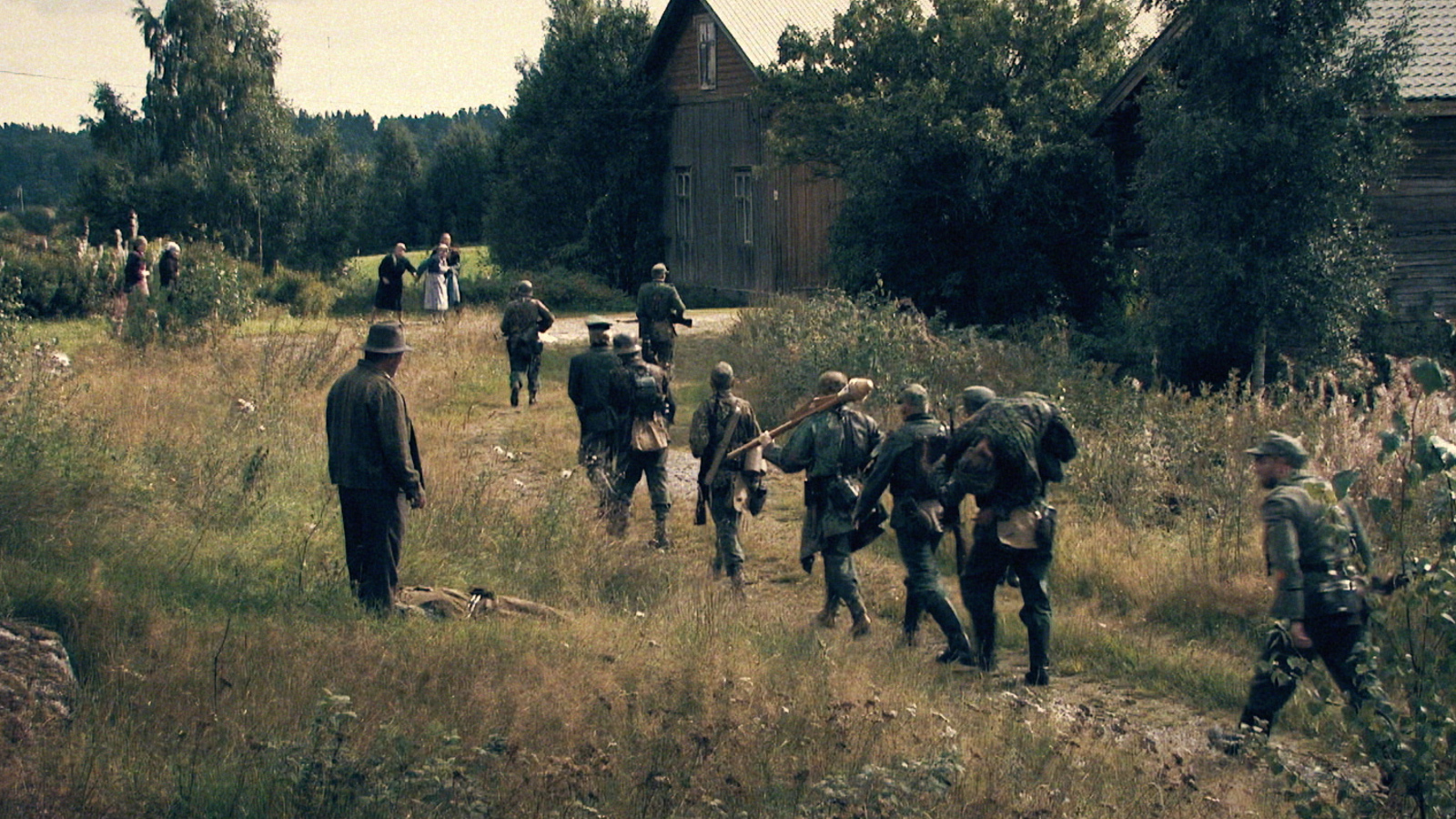 Kuvassa on sotilaita kävelemässä kohti kylää, jossa on harmaita rakennuksia molemmin puolin pientä tietä.   