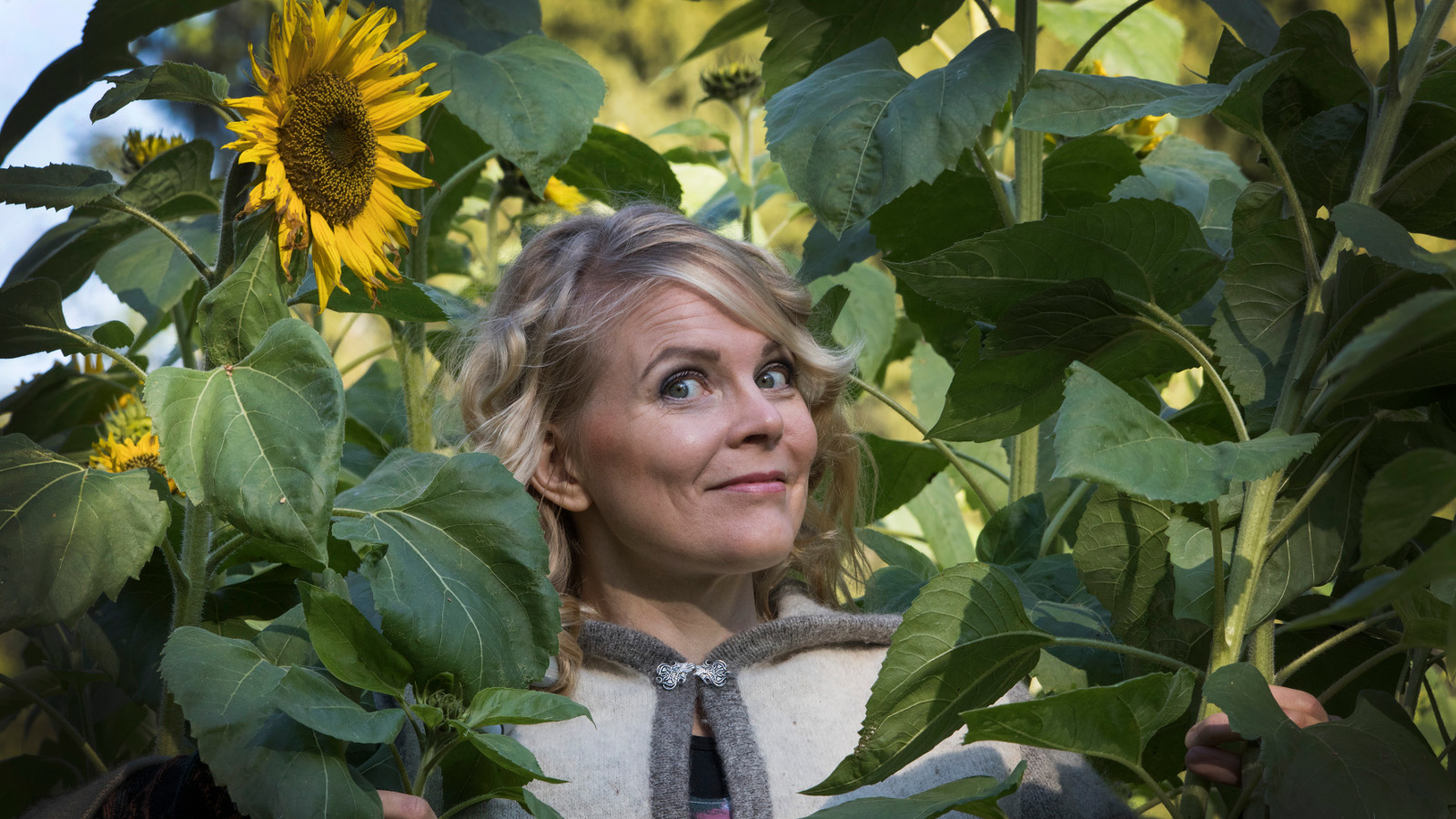 Kuvassa Heli Laaksonen on viherkasvien keskellä ja hänen takanaan on yksi auringonkukka. Kuva on puolivartalokuva.