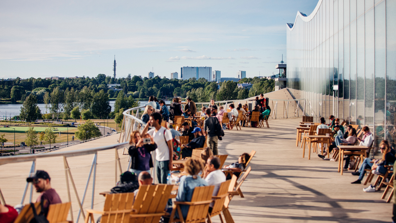 Kuvassa on ihmisiä viettämässä kesäpäivää Altaan terassilla lepotuoleissa ja takana on seinää.  Taustalla näkyy kaupunkinäkymää Helsingistä.