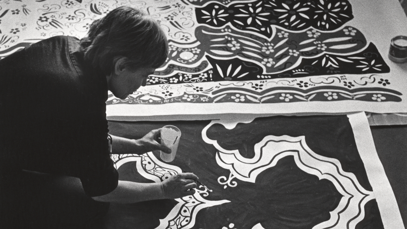 Kuvassa on Maija Isola lattialla kumartuneena maalaamaan kankaan kuvioita.  Kuva on musta-valkoinen.