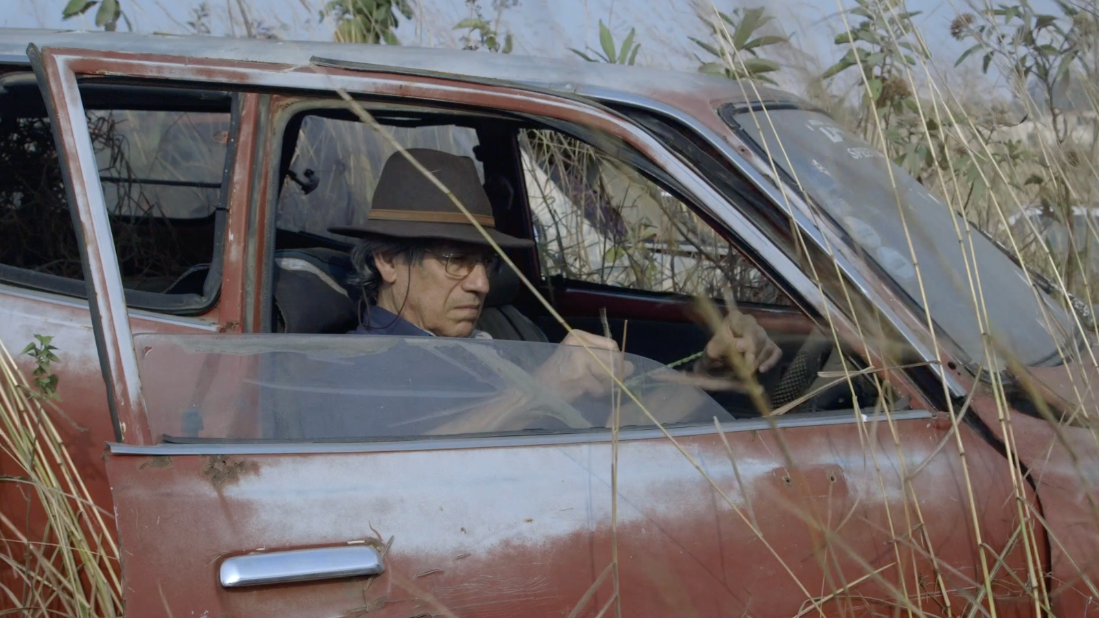 Kuvassa on vanhahko mies autossa ja auto on kuivahkossa pitkässä ruohikossa.