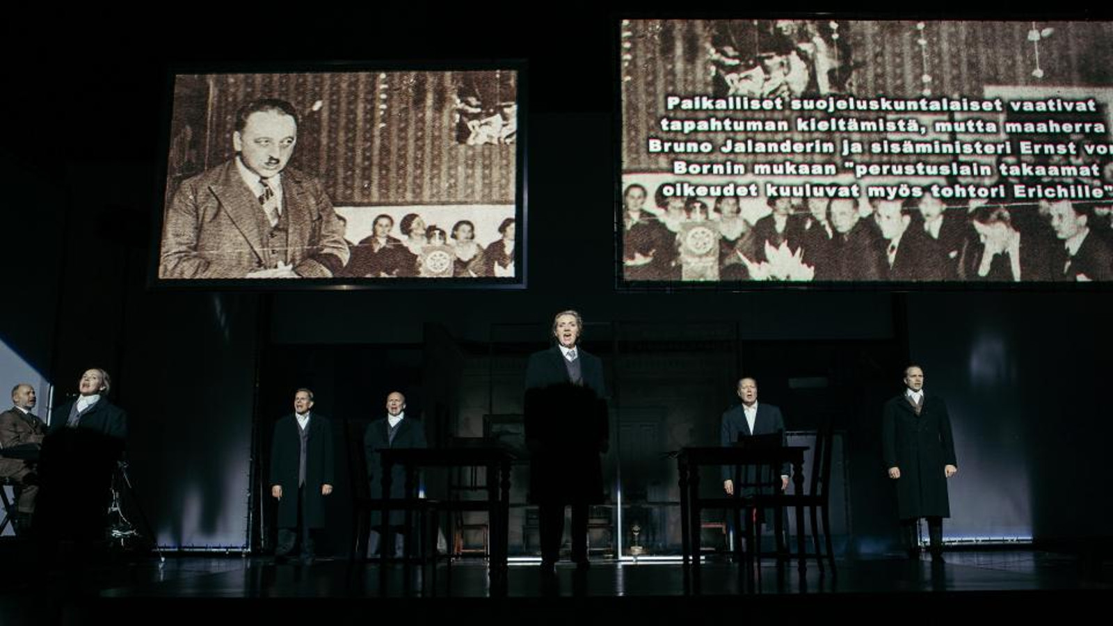 Kuvassa ovat alaosassa näyttelijät seisomassa ja laulamassa.  Yläosassa näyttötaulut, joissa oikealla on mies puhumassa ja oikealla tekstiä tapahtumasta. Kuvat ovat mustavalkoisia.