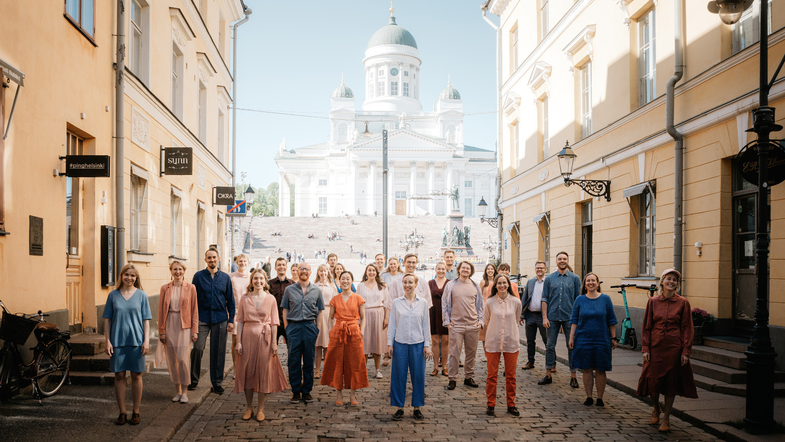 Kuvassa ovat kuorolaiset hajautuneina kadulle, jonka taustalla näkyy Helsingin tuomiokirkko. Vasemmalla ja oikealla olevat rakennukset ovat keltaisia.