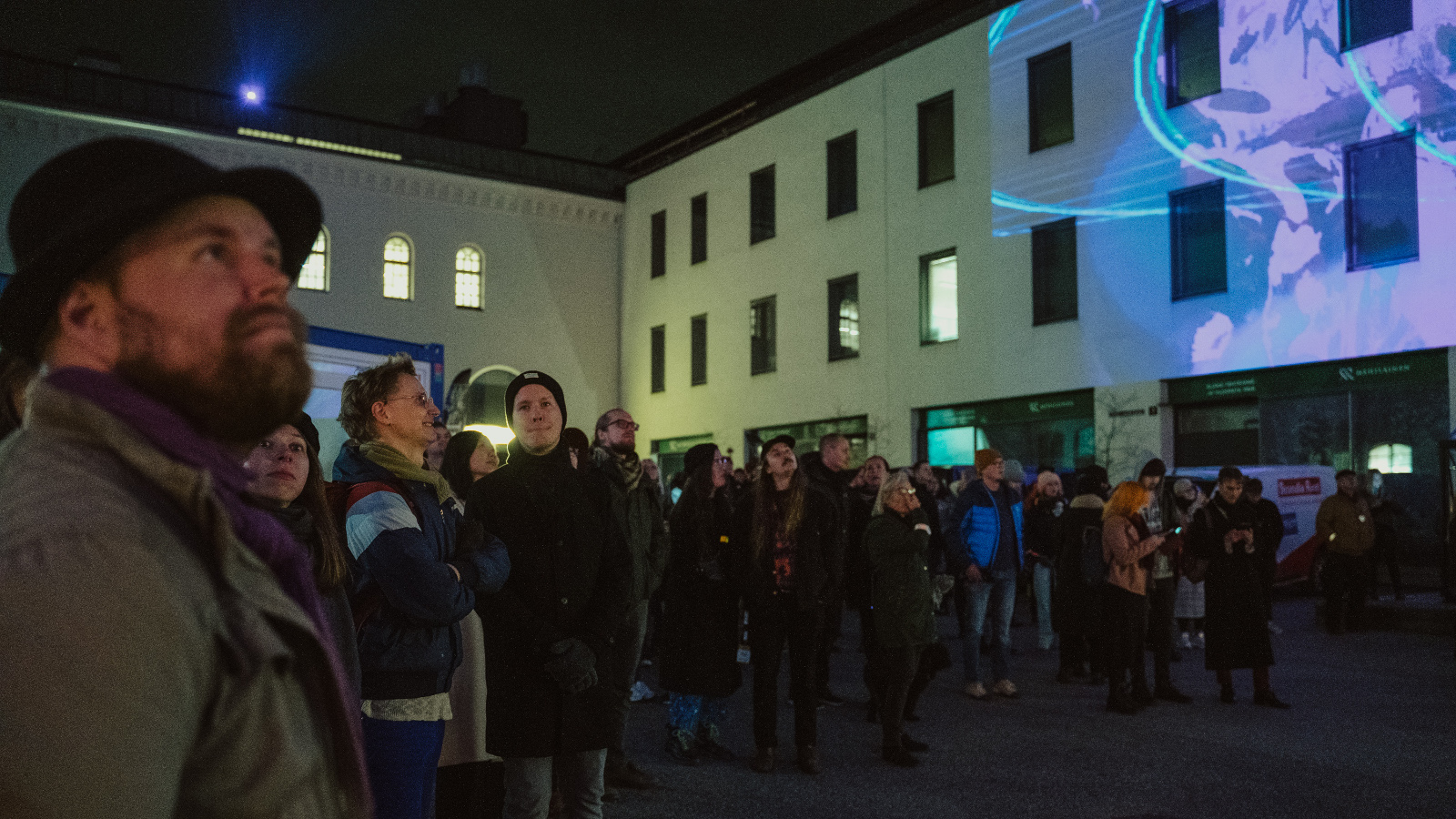 Kuvassa on kasvot eteenpäin ihmisiä katsomassa elokuvaesitystä sisäpihalla.  Kuvaa heijastuu rakennuksen vaalealle seinälle.