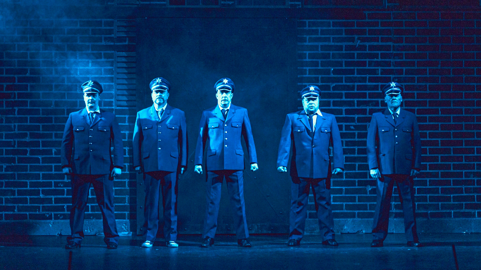 Kuvassa on viisi miestä sinisissä virkapuvuissa ja koppalakeissa.  Taustalla on tiiliseinää.  Kuvan sävy on sinertävä.