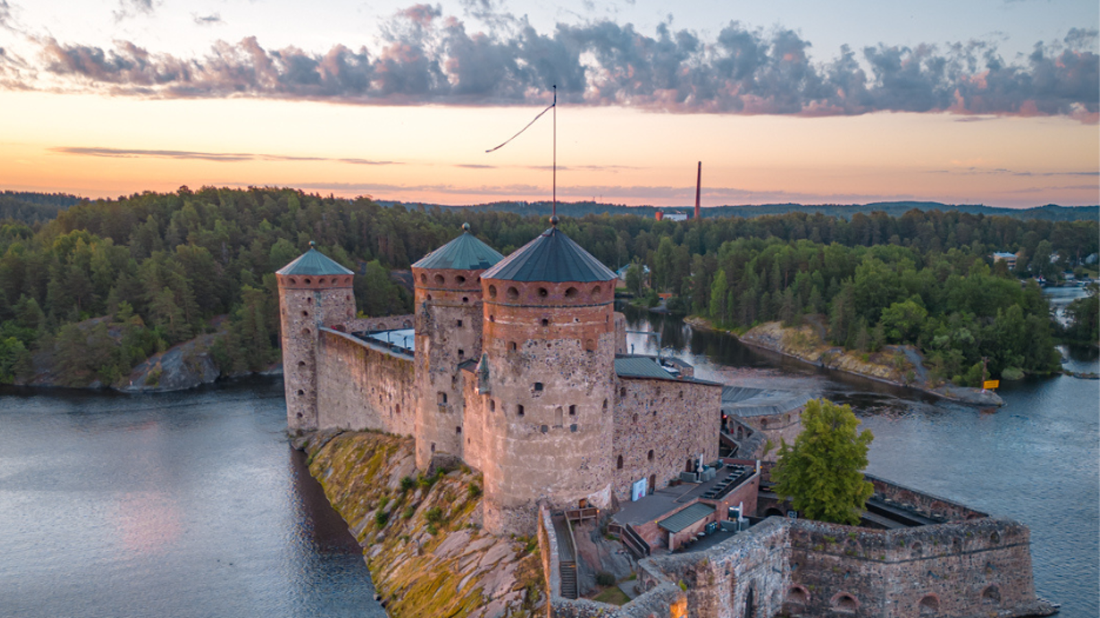 Kuvassa on Olavinlinna kuvattuna ilmasta ja yläosassa on sinertävän vaalean ruskeata pilvimuodostelmaa.